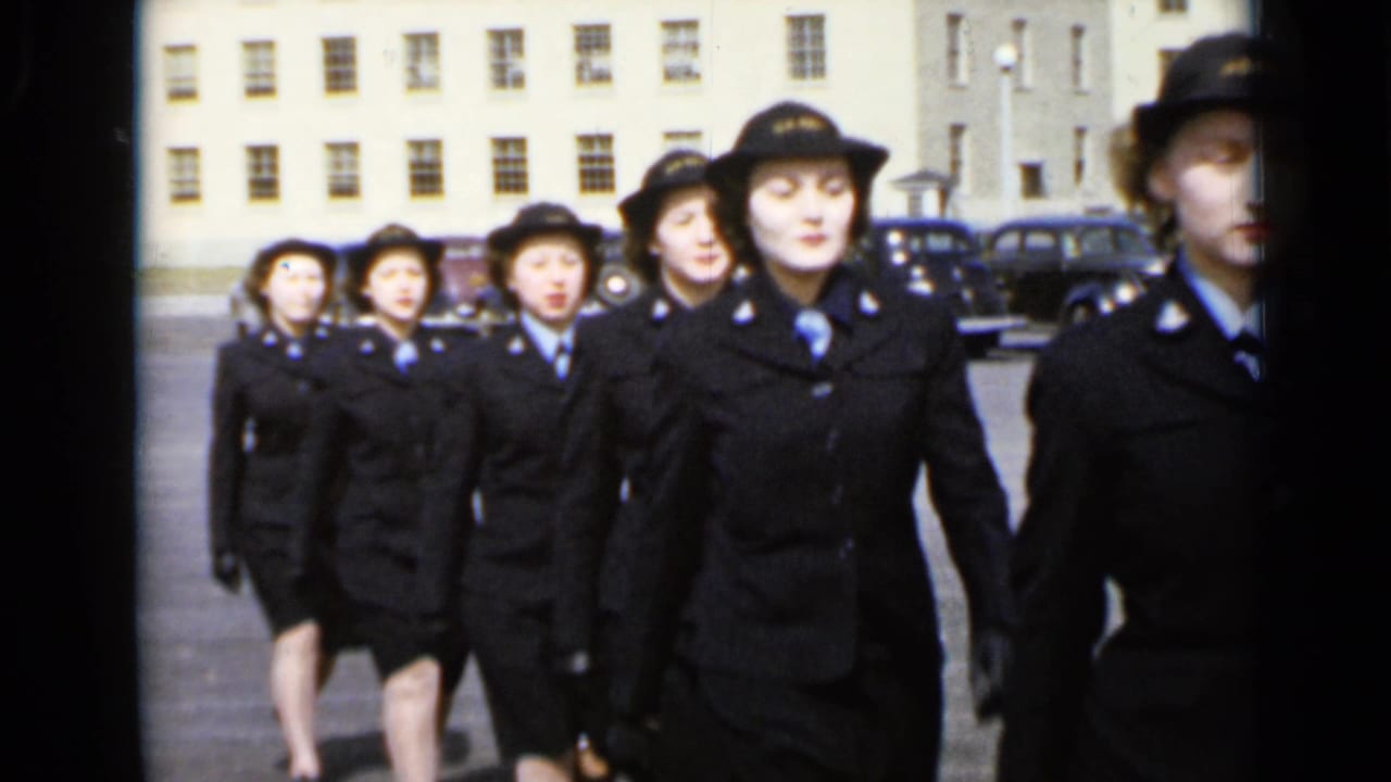 Uniformed women march toward the camera in formation outside a large building.