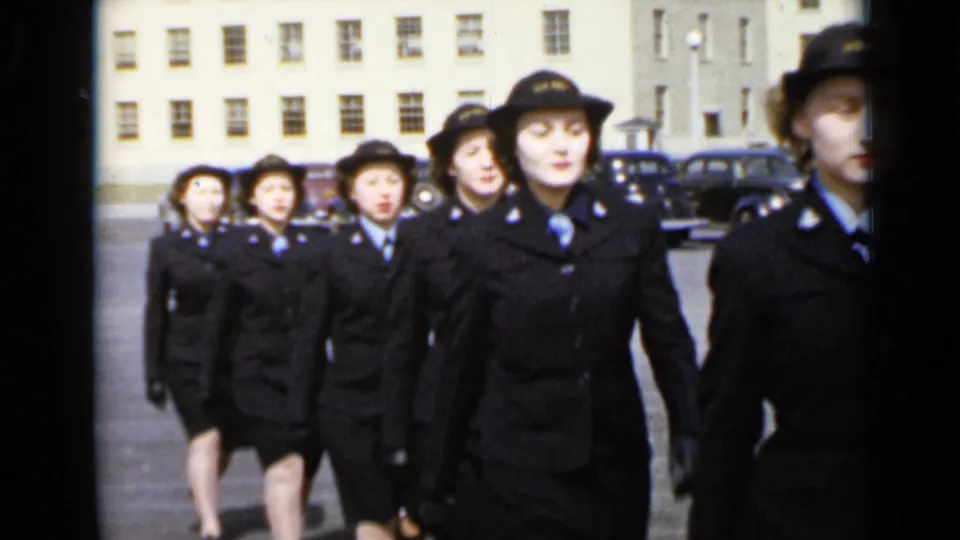 In 1947 New York, women in dark uniforms, hats, and gloves march in formation across a large paved lot with parked cars behind them.