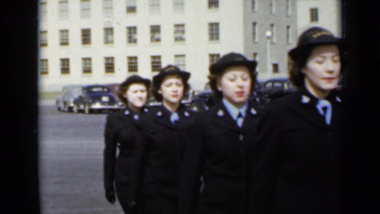 Uniformed women in hats and dark coats march in a line across the lot, with mid-century cars and a large institutional facade in the background.