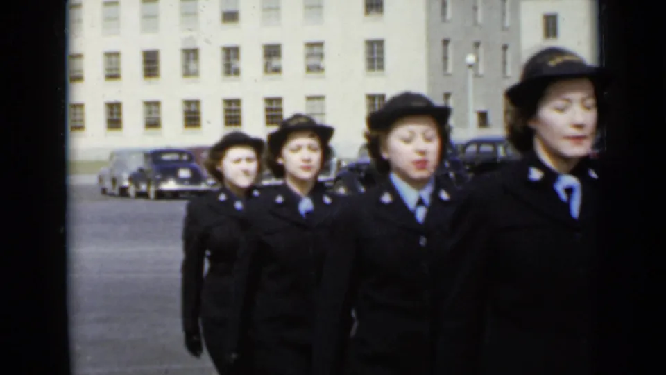 Uniformed women in hats and dark coats march in a line across the lot, with mid-century cars and a large institutional facade in the background.