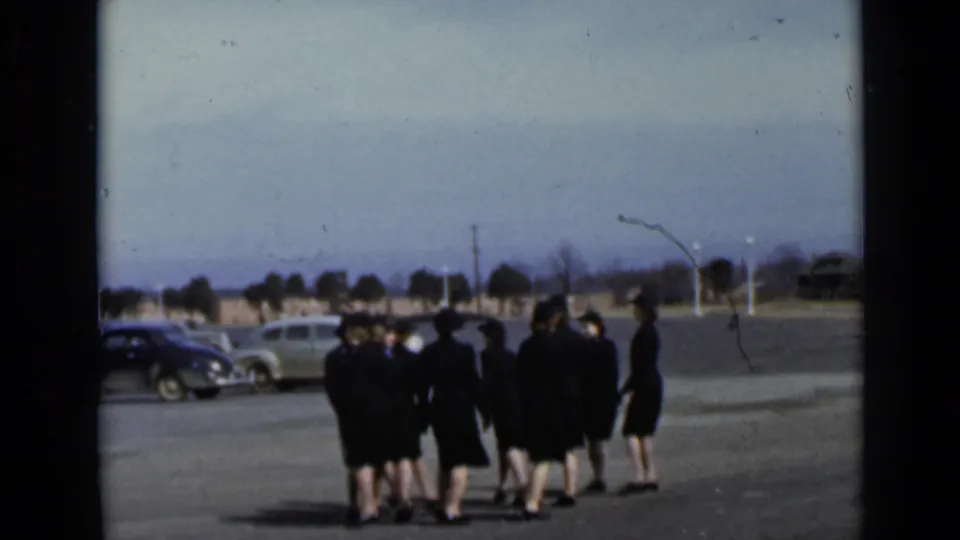 The women appear farther from the camera, grouped on the paved lot with parked cars, a road edge, and trees visible beyond them.