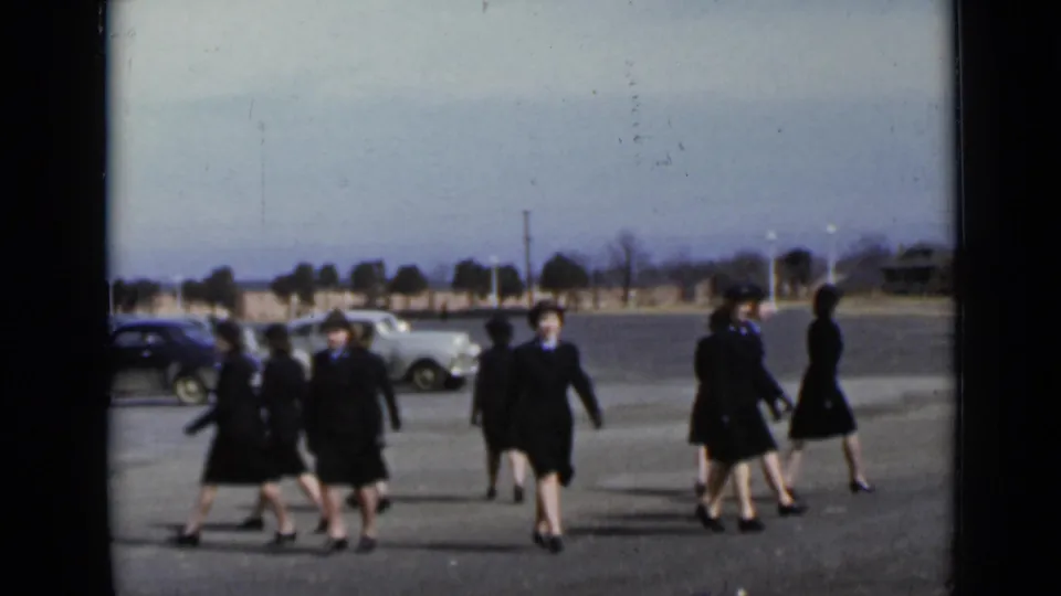 Several uniformed women cross and turn within the open lot, with vintage cars and a low tree line behind them in the New York archival footage.
