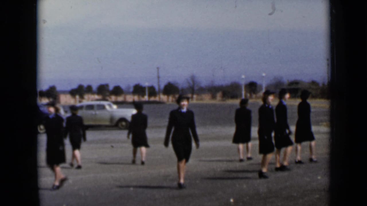 Women in dark uniforms walk across a paved lot with vintage cars in the background.