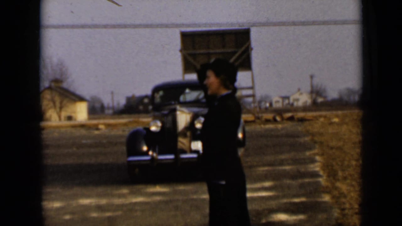 A woman in a dark uniform stands near a vintage car and a large building.