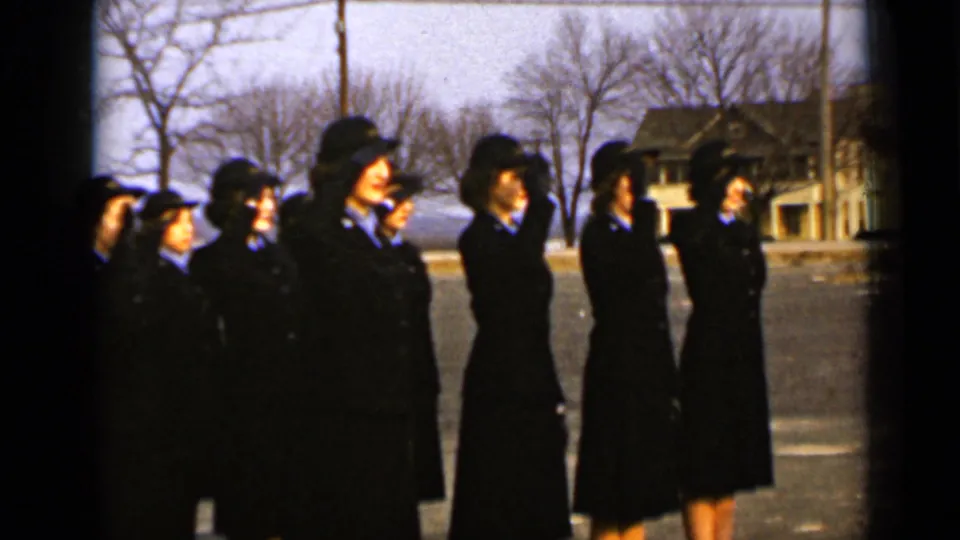 Uniformed women stand shoulder to shoulder outdoors in New York, with bare trees, a road or paved area, and houses visible in the background.