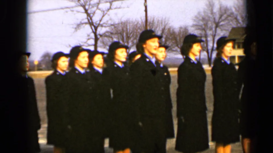 Women in matching dark uniforms and hats stand in a close formation, facing toward the right, with trees and houses behind them.