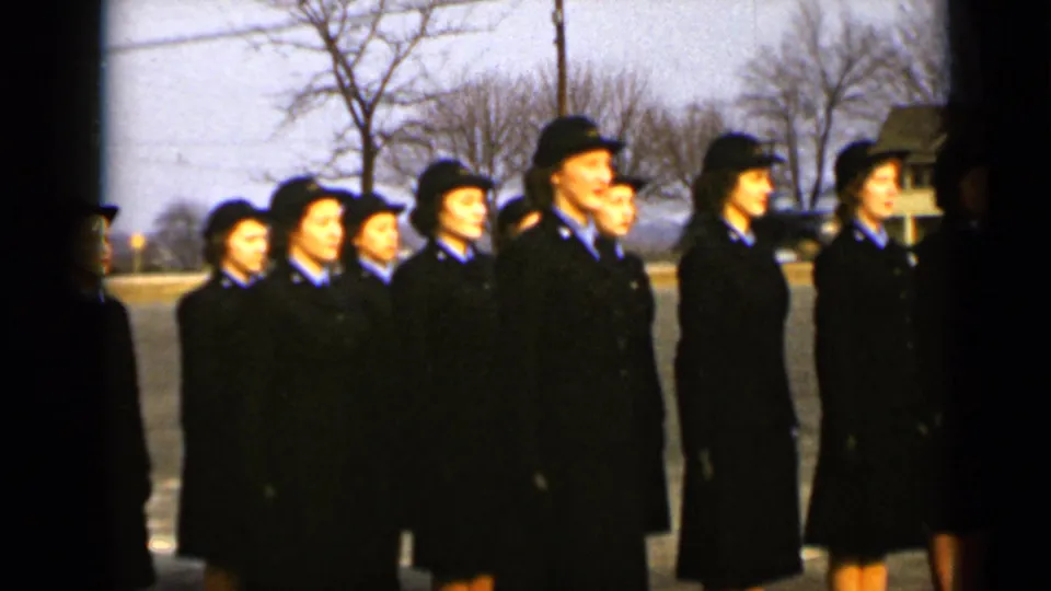 Uniformed women in dark coats and blue collars stand in formation on a New York street in 1947, with bare trees behind them.