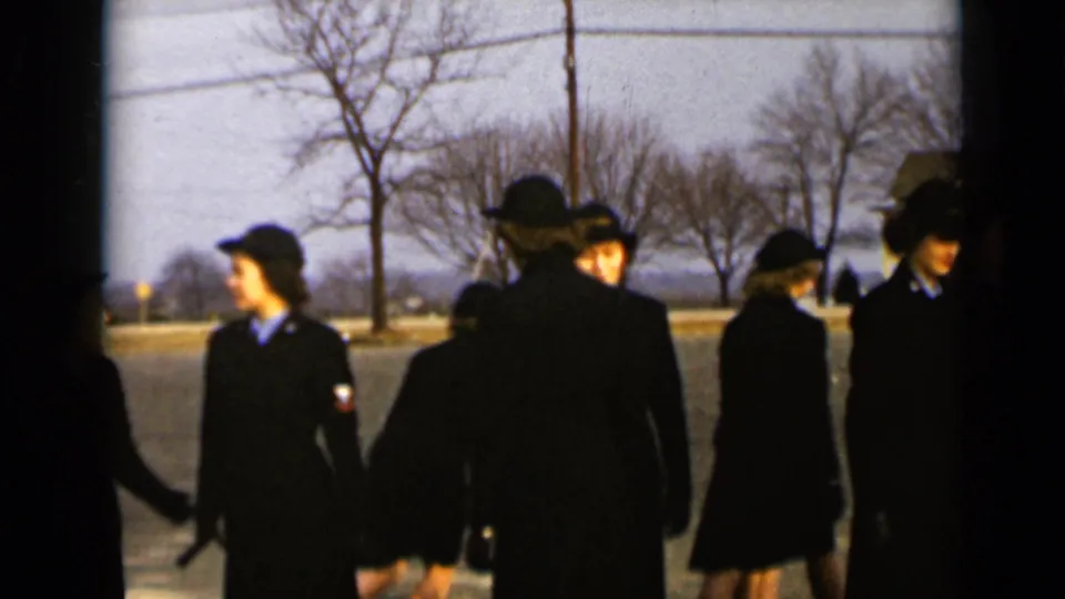 Several uniformed women in dark coats and hats shift position on a New York street, with leafless trees and houses in the background.