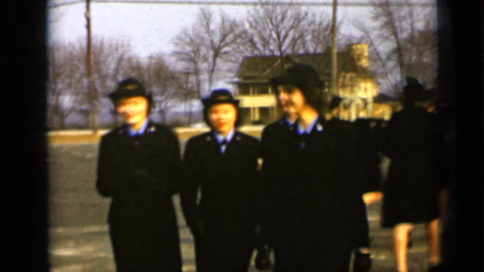 Three women in matching dark uniforms face the camera in 1947 New York footage, with other figures and a residential house behind them.