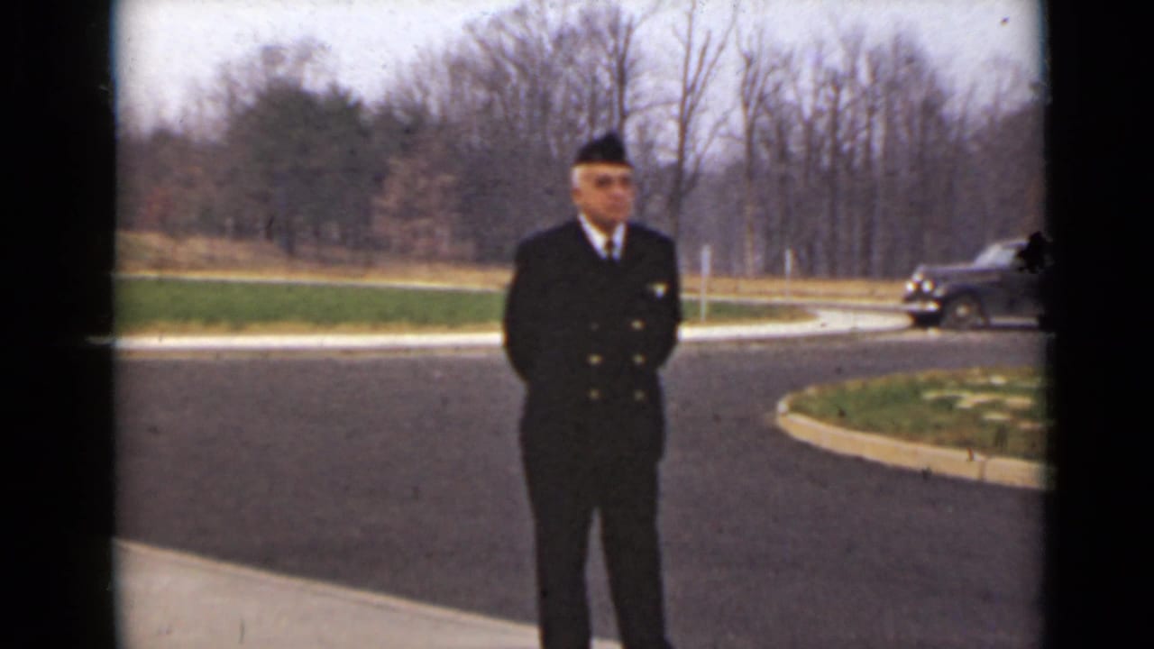 Uniformed man standing on a paved road with bare trees and a dark car in the background.