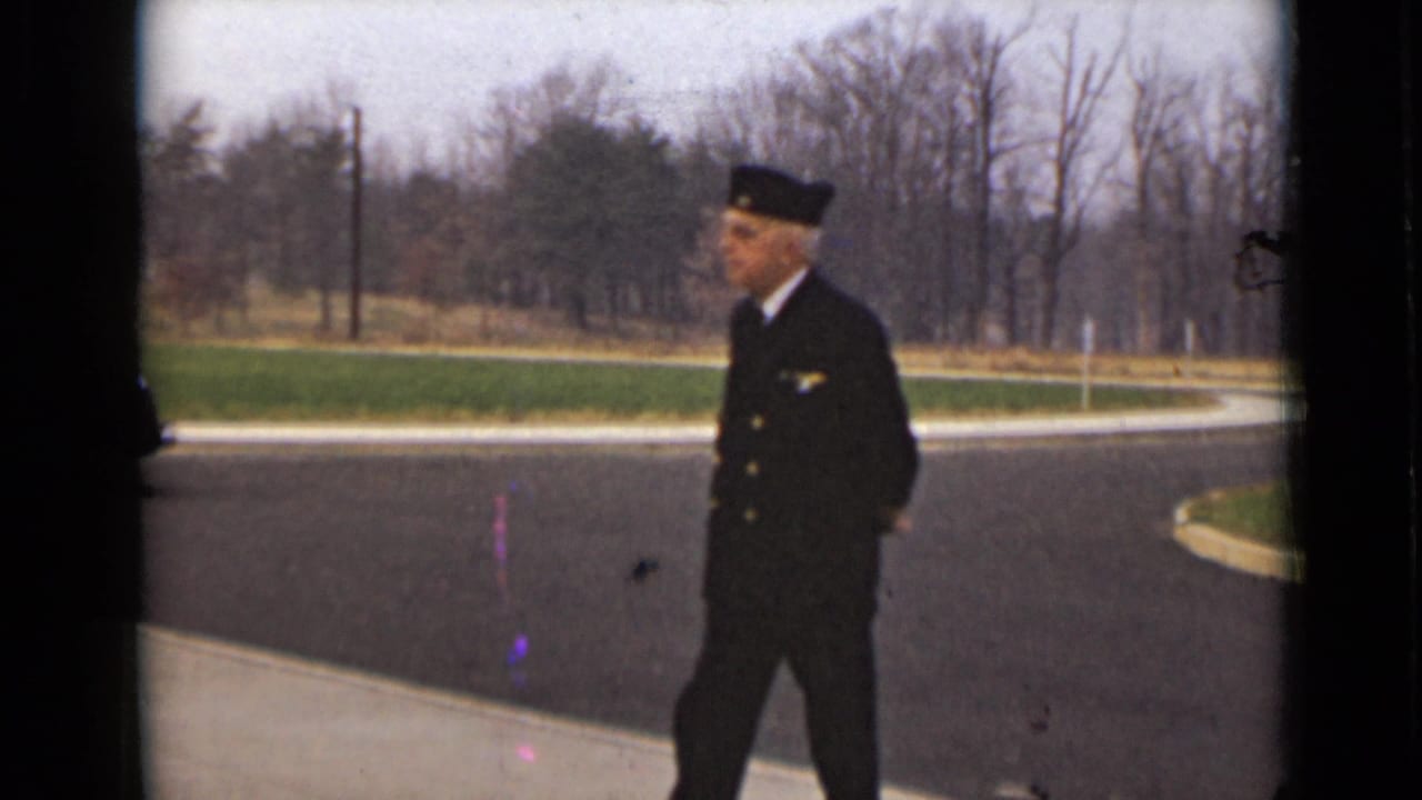 Uniformed man walking across a paved road near grass and bare trees.