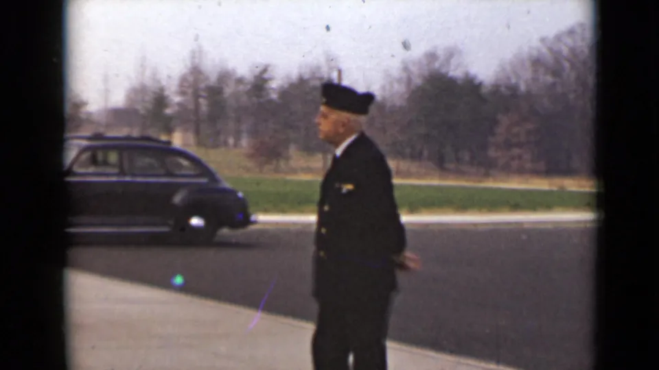 A uniformed man passes a dark rounded sedan parked along the road, with grass and bare trees visible in the background.
