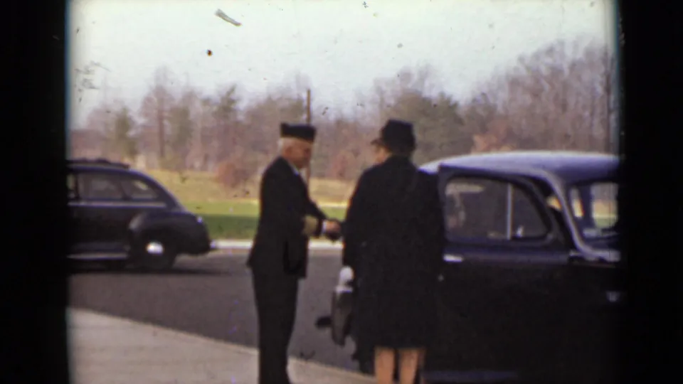 In the 1947 New York scene, the uniformed man assists a woman in a dark coat and hat beside a dark car near another parked sedan.