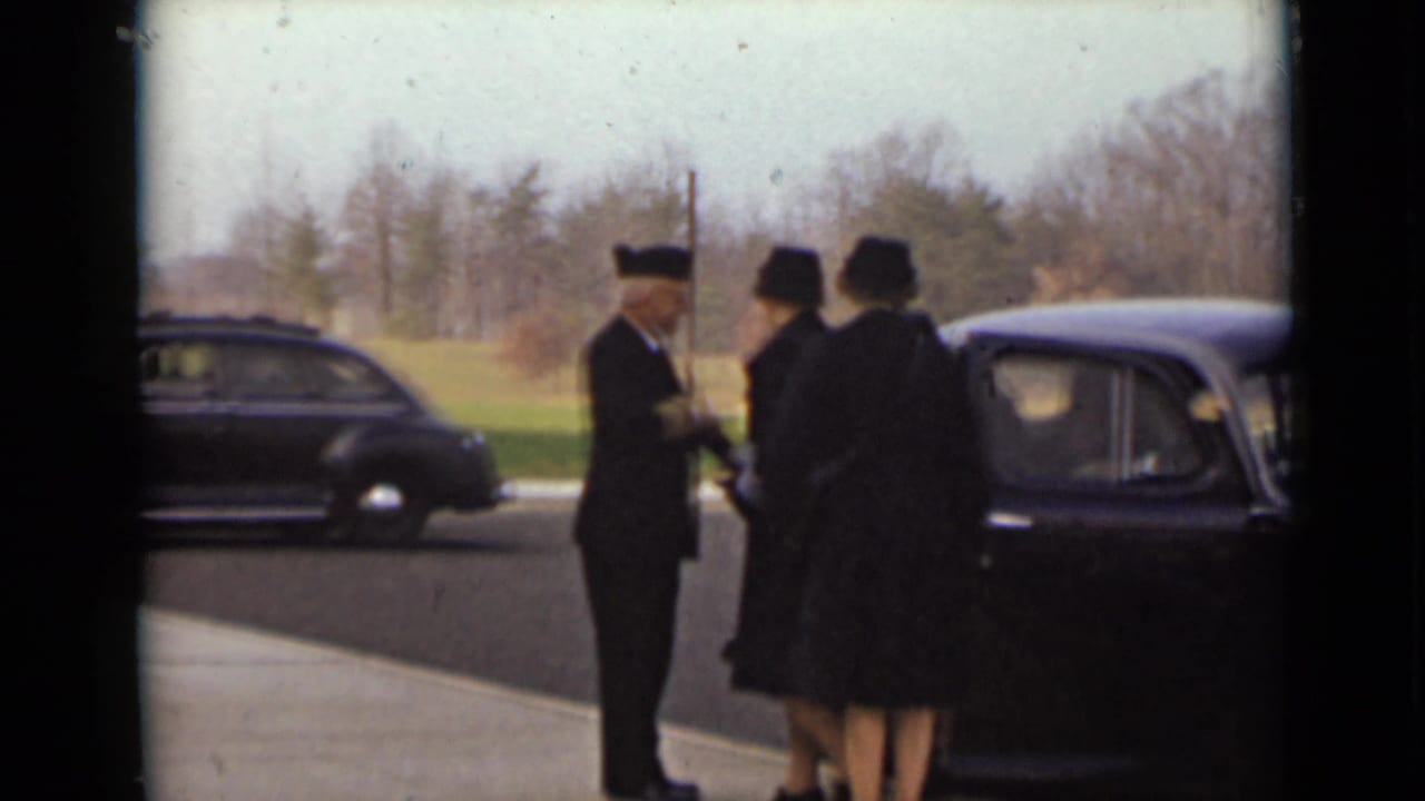 Uniformed man shakes hands with women in dark coats beside a black car.