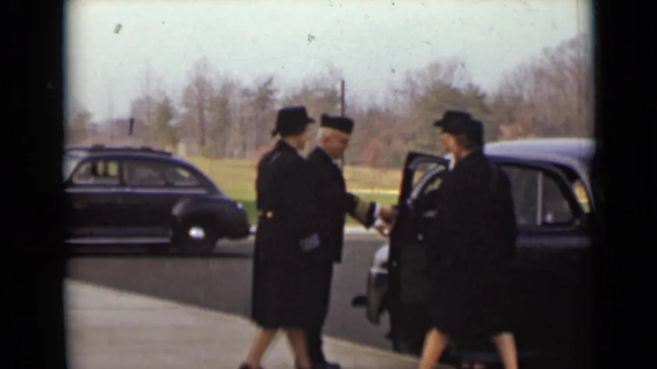 Women in dark coats and hats move around a black car while a uniformed man stands near the open door in New York, 1947.