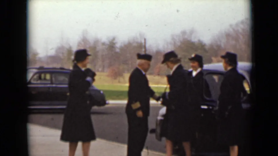 A uniformed man shakes hands with a dark-coated man while women stand nearby beside parked cars in the 1947 New York scene.