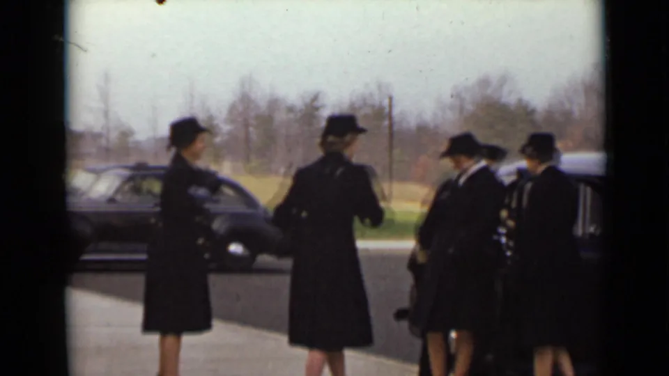 Women in black coats and hats stand beside a dark sedan on a paved roadside, with another car and bare trees in the background.