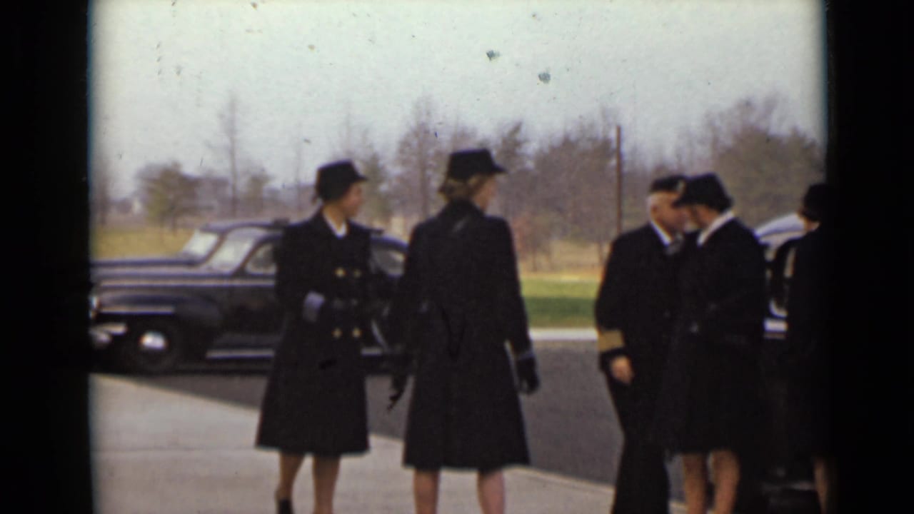 Uniformed women and men stand near a black car in an outdoor lot.