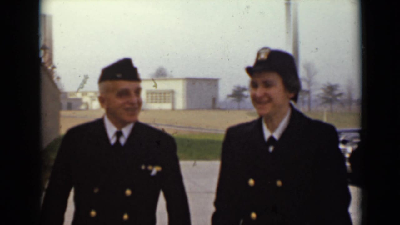 Two uniformed adults walk together past a paved lot and low building.