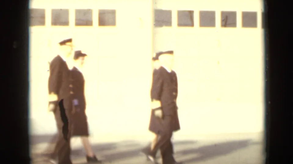 Uniformed adults pass in front of bright bay doors, with overexposed 8mm sunlight washing part of the frame.