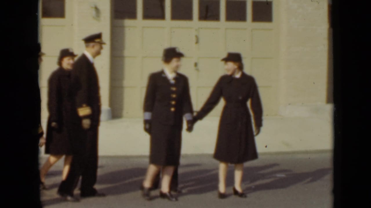 Uniformed adults walk past a pale garage door in bright sunlight.