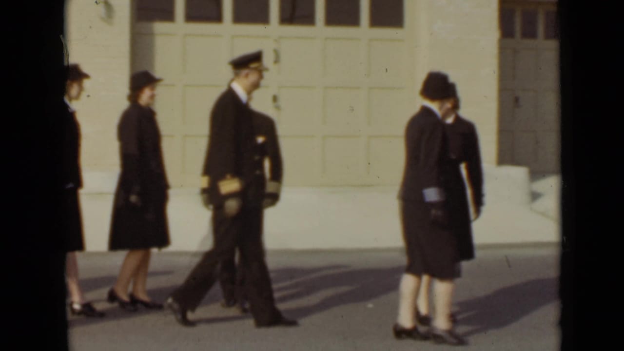 A man in a naval-style uniform walks while uniformed women pass nearby.