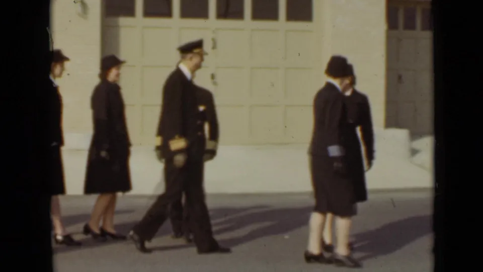 In 1947 New York, a man with gold sleeve bands walks near women in dark uniforms outside a pale garage door.