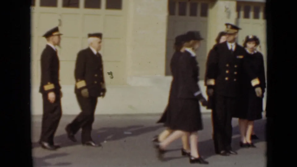 In 1947 New York, uniformed adults cluster and walk near a white garage-style door, with faces visible in soft 8mm color.