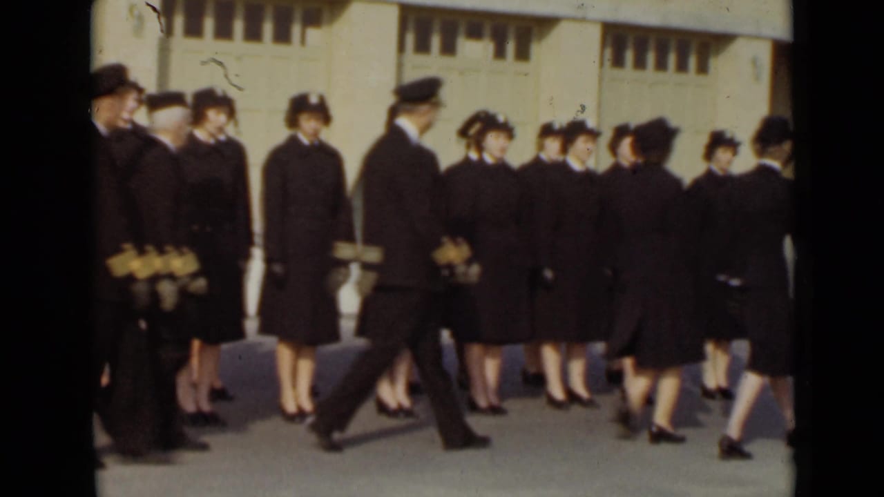 Uniformed men and women stand and begin marching outside a large building.