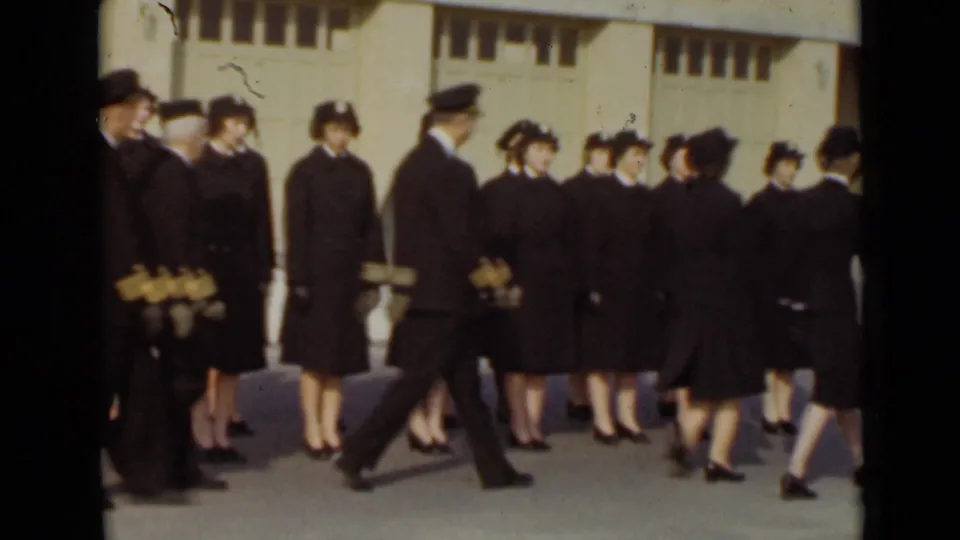 In 1947 New York, uniformed men and women in dark coats and hats gather and move in formation outside a large building.