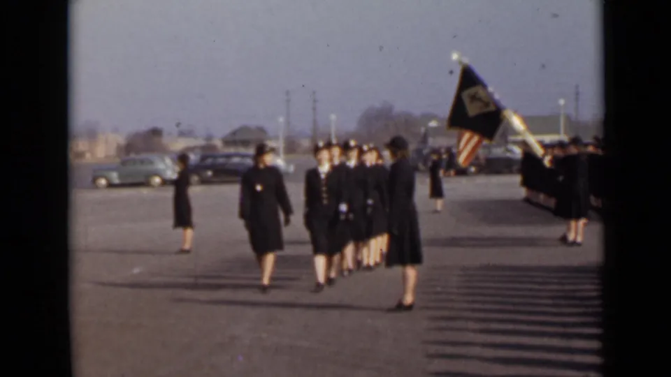 A flag bearer and uniformed participants march across an open paved area with parked cars in the background in 1947 New York.
