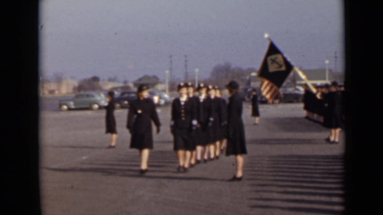 Women in dark uniforms march across a paved lot while a flag bearer stands nearby.