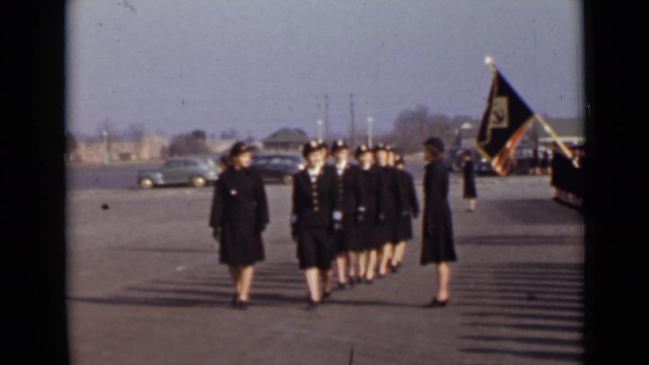 Uniformed women march toward the camera as a flag bearer stands at the right.