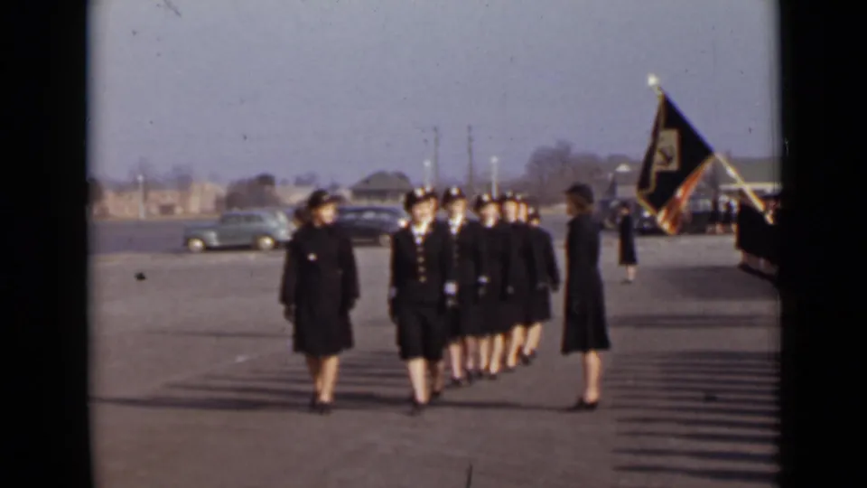 Women cadets in dark uniforms advance in formation on a paved New York lot, with a flag bearer and vintage cars behind them.