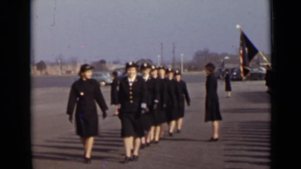 Uniformed women continue marching toward the camera on a paved lot in New York, with a dark flag and mid-century cars in the background.