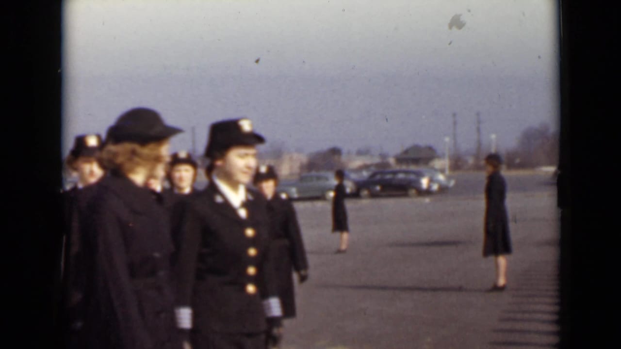 Women in dark uniforms march across a paved parade ground with cars in the distance.