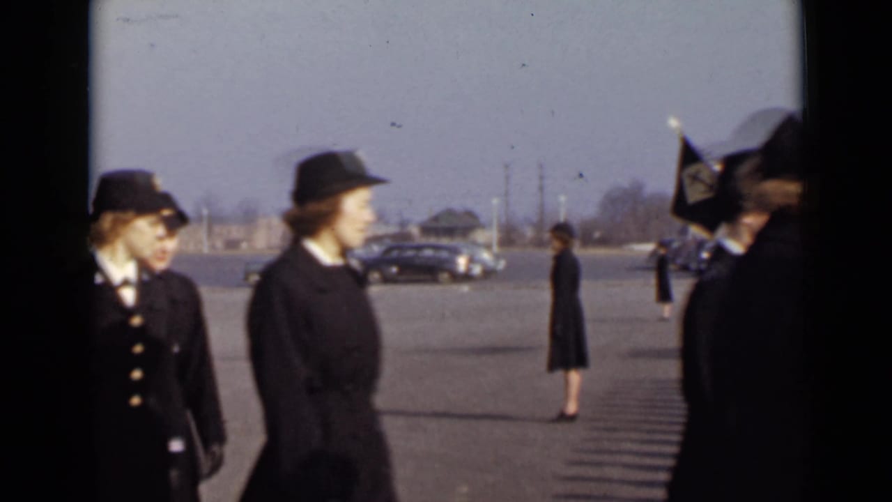 Women in dark service-style uniforms move through the foreground while flags, cars, and a lone uniformed figure remain behind them.