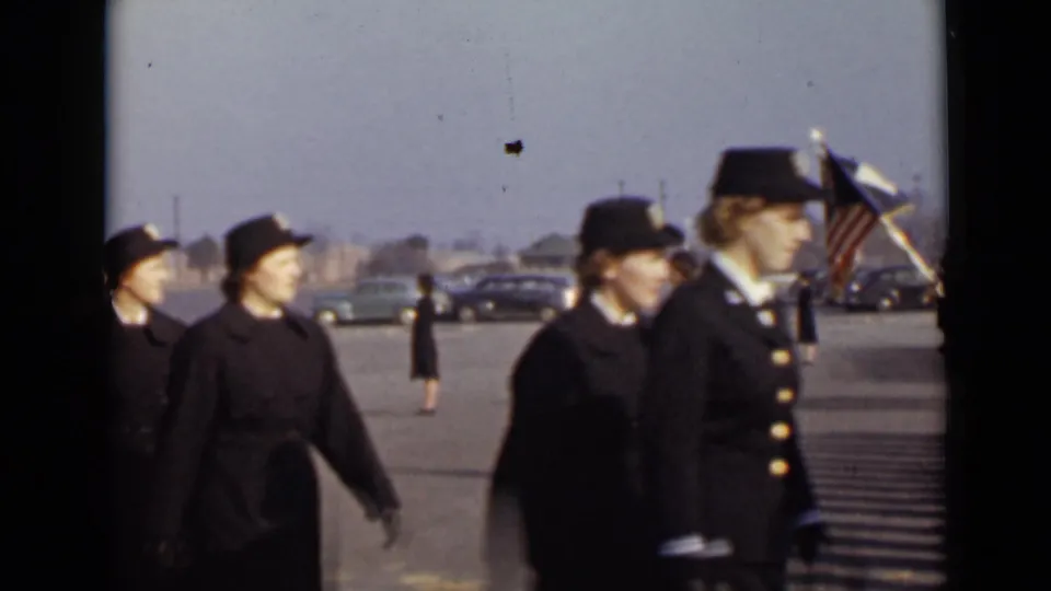 1940s home-movie frame of women in dark skirted uniforms marching in line, with flags and parked mid-century cars in the background.