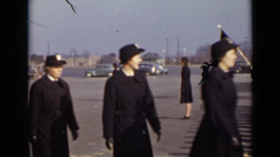 Women in dark uniforms march through the foreground in New York footage dated 1947, with cars and a standing figure visible beyond them.