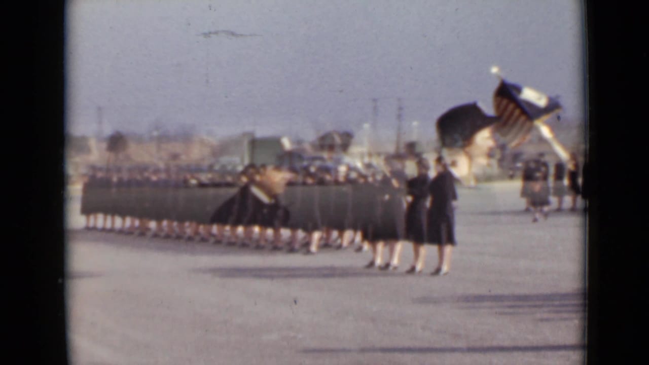 Uniformed women march in formation across a paved lot while flag bearers stand near the front.
