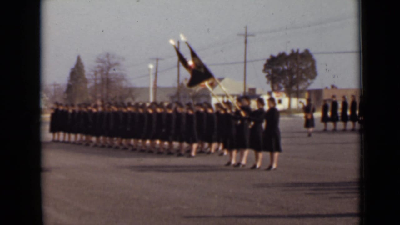 Rows of uniformed women stand or march in formation with flags raised at the front.