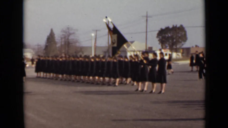 The 8mm frame shows uniformed women marching in formation in New York, with a large flag, utility poles, and low buildings behind them.