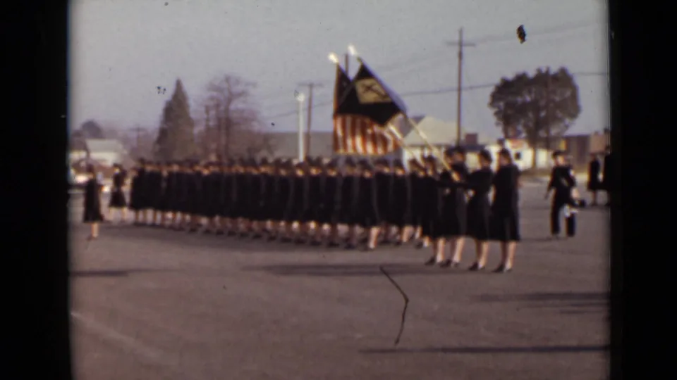 1947 New York frame of women in matching dark uniforms marching in rows, with an American flag and another flag visible at the front.