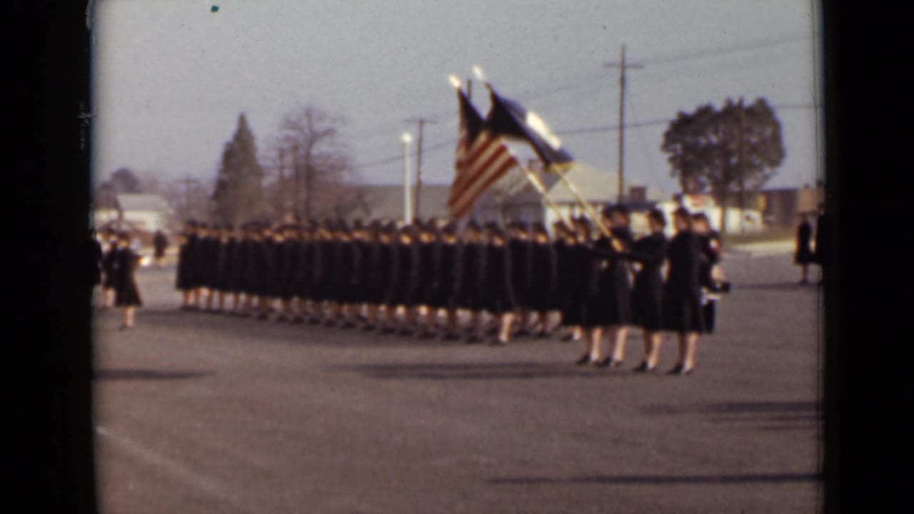 Uniformed women stand in formation with American and service flags on a paved outdoor area.