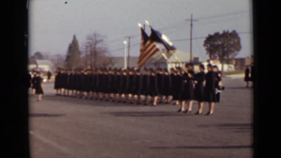 In 1947 New York, women in dark uniforms line up for a military-style ceremony while flags are held at the front.