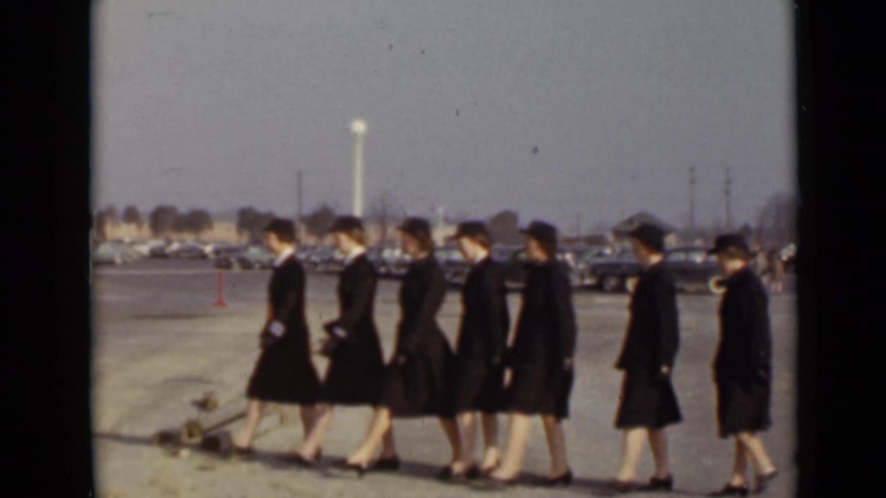Women in dark uniforms march in a line across a paved lot with parked cars behind them.