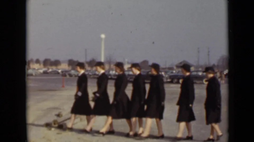 Uniformed women march in step across an outdoor paved area in New York, with parked period cars visible in the background.
