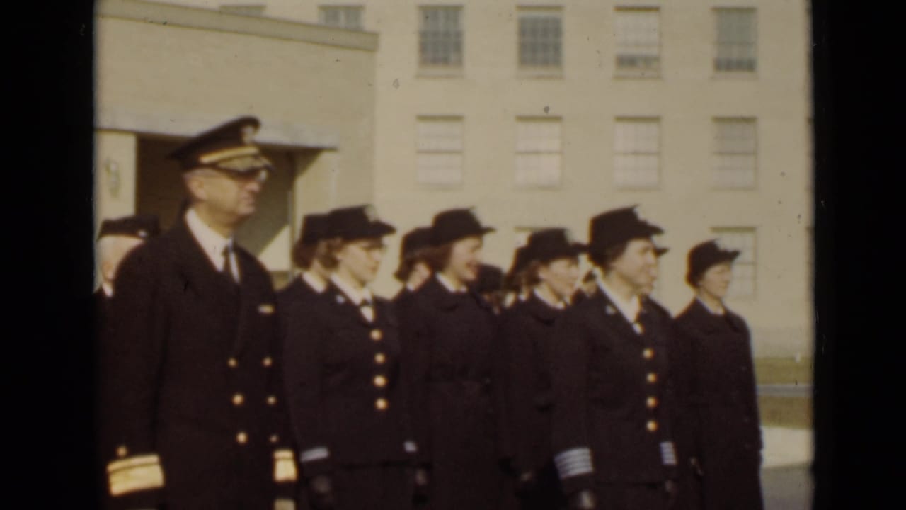 Uniformed women and a male officer stand at attention outside a light-colored building.