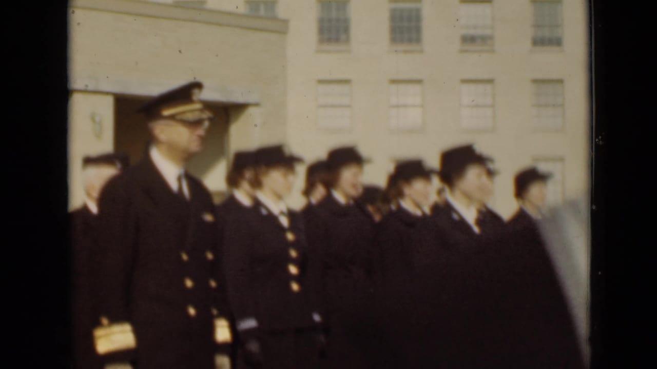 A row of uniformed women stands beside a male officer in front of a large building.