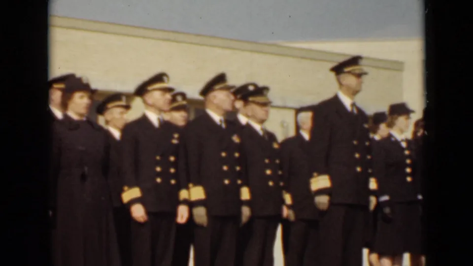 The 1947 New York home-movie frame shows officers and women in dark uniforms standing at attention outside a pale building.